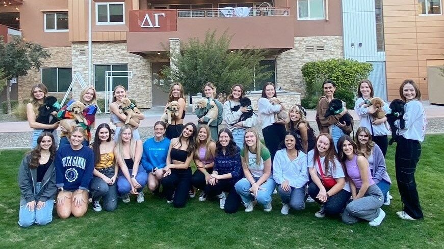 A large group of chapter members pose in front of the house. 10 of the members are holding guide dog puppies.