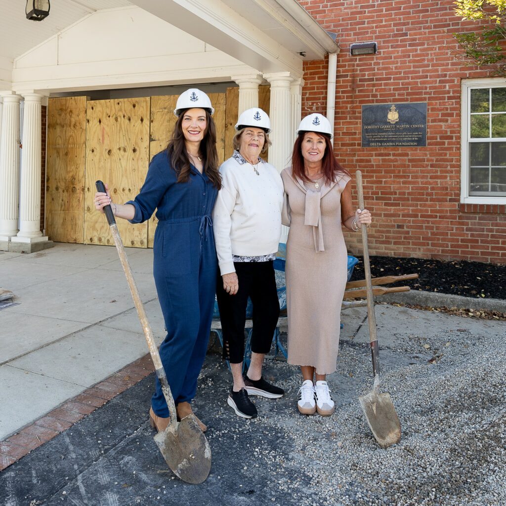 Leslie, Judy, and Tracey stand outside of Executive Offices. There is a pile of gravel and wheelbarrow next to them. They are all wearing hard hats with the DG logo on them and holding shovels.